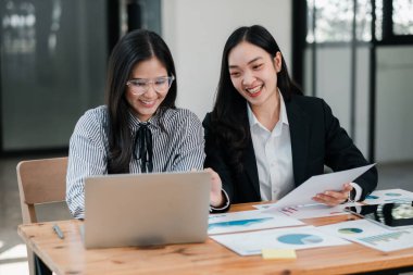 İki iş kadını, bir dizüstü bilgisayar ve belgelerle bir masada çalışmayı, profesyonel bir ortamda takım çalışması ve işbirliği sergilemeyi tartışıyor..