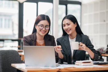 İki iş kadını, parlak ve modern ofis ortamında dizüstü bilgisayar ve kahveyle bir masada çalışmayı tartışıyor..