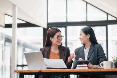 Modern bir ofis ortamında iki iş kadını, belgeler ve tabletle çalışma, takım çalışması ve işbirliği hakkında tartışıyorlar..