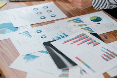 Ahşap bir masa üzerinde renkli tablolar ve grafiklerle finansal belgeleri analiz eden bir iş adamı, veri analizi ve stratejiyi ön plana çıkarıyor..