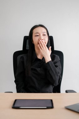 Siyah gömlekli profesyonel bir kadın, elinde tabletle masada otururken esniyor, kurumsal bir ortamda yorgunluk ve bıkkınlık taşıyor..