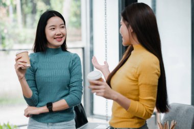 İki kadın, gündelik kazaklar içinde ellerinde kahve fincanları, parlak, modern ofis ortamında sohbet ediyorlar..
