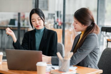 Modern bir ofiste iki iş kadını, masada kahve fincanları olan bir dizüstü bilgisayar kullanarak işi tartışıyor..