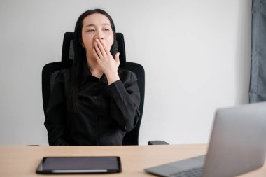 İş kadını, modern ofis ortamında dizüstü bilgisayar ve tabletle çevrili masasında esniyor..