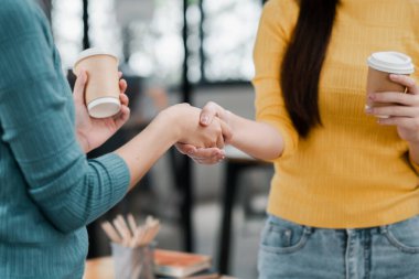 İki kadın bir ofiste el sıkışıyor, takım çalışmasını ve işbirliğini sembolize ediyor, ellerinde kahve fincanları var..