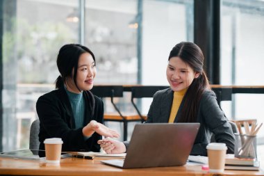 Çağdaş bir ofis ortamında birlikte çalışan iki profesyonel kadın masada kahve fincanlarıyla dizüstü bilgisayarın üzerinde fikir alışverişinde bulunuyorlar..