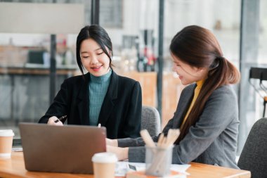 Modern bir ofiste çalışan iki genç kadın bir dizüstü bilgisayarla iş tartışıyor, takım çalışması ve işbirliği sergiliyorlar..