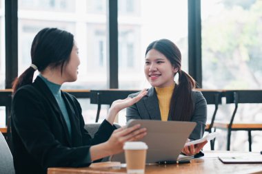 Modern ofis ortamında iki iş kadını dizüstü bilgisayarlar ve kahveyle çalışma, takım çalışmasını ve profesyonel etkileşimi tartışıyor..