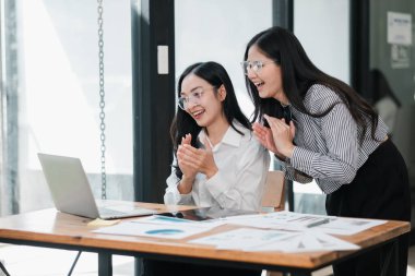 Modern ofis ortamında iki hevesli kadın, masadaki belgelerle birlikte bir dizüstü bilgisayarla çalışırken başarıyı kutluyorlar..