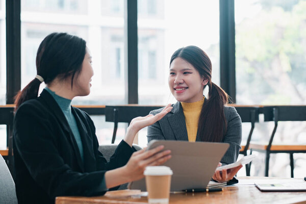 Две деловые женщины в современном офисе, обсуждающие работу с ноутбуками и кофе, подчеркивающие командную работу и профессиональное взаимодействие.