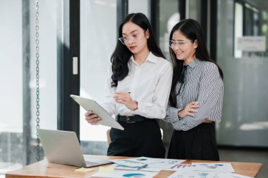 Parlak bir ofis ortamında tablet üzerinde çalışan iki profesyonel kadın. Etrafı çizelge ve dizüstü bilgisayarla çevrili..