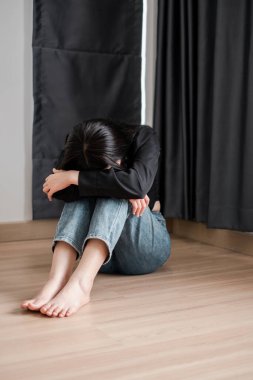 Genç bir kadın, minimalist bir odada, dizlerini kucaklayarak, yerde oturuyor..