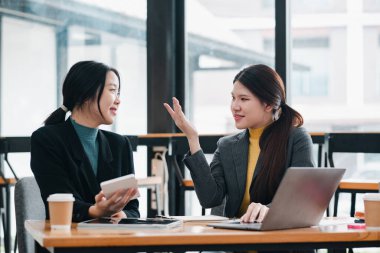 Modern ofis ortamında iki kadın, iş stratejilerini bir dizüstü bilgisayar ve akıllı telefonla tartışıyorlar..