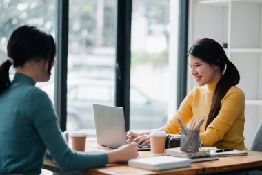 İki kadın parlak bir ofiste birlikte çalışıyor, dizüstü bilgisayar kullanıyor ve kahveden zevk alıyor, takım çalışmasını ve modern iş ortaklığını sembolize ediyor.