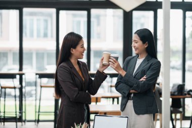 Resmi giyinmiş iki iş kadını aydınlık ve modern ofis ortamında kahve molası veriyorlar..