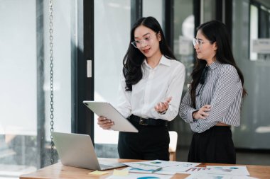 İki kadın modern ofis ortamında iş stratejilerini tartışıyor, tablet kullanıyor ve belgeleri inceliyor..
