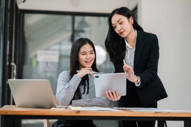 Modern bir ofiste çalışan iki iş kadını, iş yerinde takım çalışması ve teknoloji sergileyen bir tablet ve dizüstü bilgisayar kullanarak işbirliği yapıyorlar..