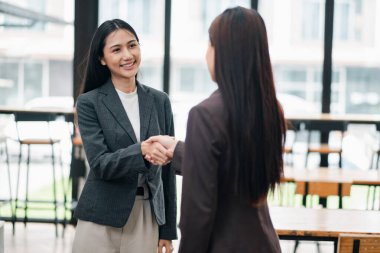 Resmi giyinmiş iki iş kadını, geniş pencereli, modern bir ofiste el sıkışıyor..