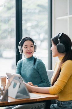 İki kadın kulaklık takıyor, sohbet ediyor ve büyük pencereli, modern bir kafede müzik dinliyor..