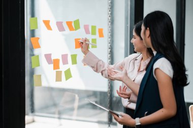 Cam bir duvara yapışkan notlar kullanarak çağdaş ofis ortamında yaratıcılığı ve takım çalışmasını teşvik eden iki profesyonel beyin fırtınası yapıyorlar..