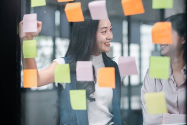 Çağdaş bir ofiste fikir üretmek için cam bir duvara yapışkan notlar yapıştırarak beyin fırtınası yapan iki meslektaşım..