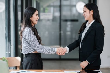 Two businesswomen in formal attire shaking hands in a bright, modern office environment, symbolizing partnership and collaboration.