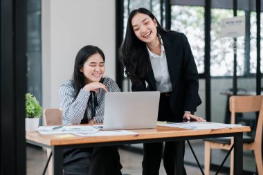 Modern ofis ortamında iki Asyalı iş kadını, ahşap bir masanın üstünde gülümsüyor ve iş birliği yapıyorlar..