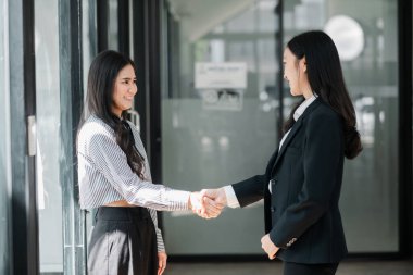 Parlak, modern ofis ortamında resmi giyinmiş iki iş kadını el sıkışıyor..