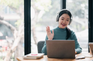 Ev ofisinden bilgisayar ve kulaklık kullanan, gülümseyen ve el sallayan bir kadın..