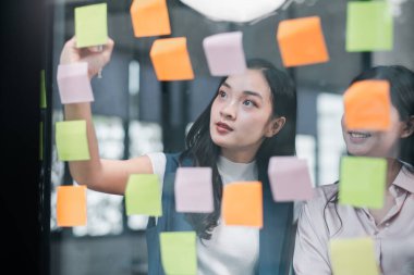 Modern ofis ortamında cam bir duvara renkli notlar yapıştıran iki genç profesyonel beyin fırtınası..