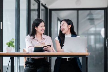 Çağdaş bir ofiste iş birliği için dizüstü bilgisayar ve not defterlerini kullanan iki kadın üretken bir tartışmaya giriştiler..