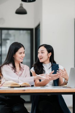 Modern bir ofis ortamında iş birliği yapan iki kadın, iş stratejilerini dizüstü bilgisayarlar ve belgelerle tartışıyor..