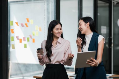 İki iş kadını renkli yapışkan notlarla çevrili çağdaş bir ofiste fikirleri tartışıyor, ellerinde bir tablet ve kahve tutuyorlar..