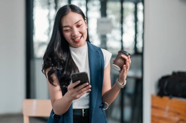 Gülümseyen genç bir kadın elinde kahve ve akıllı telefon ile modern ofis ortamında eğleniyor..