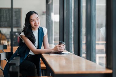 Gülümseyen kadın kahve fincanı tutuyor, modern bir kafede pencere kenarında oturuyor, rahatlatıcı bir anın tadını çıkarıyor..