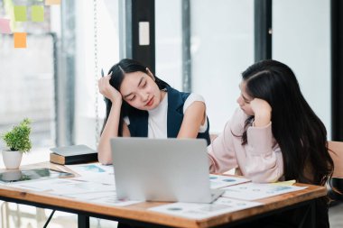 İki genç kadın, bir masa başında dizüstü bilgisayar ve belgelerle tartışırken modern ofis ortamında takım çalışması sergiliyorlar..