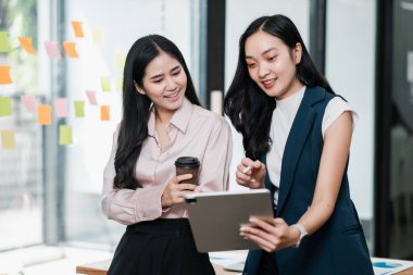 Çağdaş bir ofiste çalışan, tablet kullanan ve kahvenin tadını çıkaran iki iş kadını..