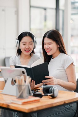 İki genç kadın bir kitabı paylaşıyor ve modern bir kafede tablet kullanıyor, gülümsüyor ve ders çalışmaktan zevk alıyorlar..