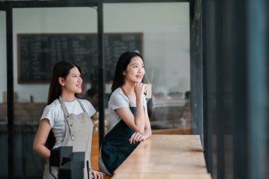 Önlüklü iki genç barmen modern bir kahve dükkanında bir pencerenin önünde durup rahatlama anının tadını çıkarıyorlar..