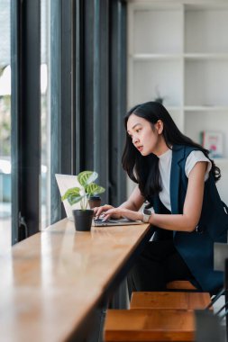 Geniş pencereli ve minimalist dekorlu parlak, modern bir ofis alanında dizüstü bilgisayar kullanan odaklanmış bir kadın..