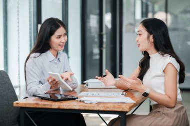 Modern ofis ortamında iş görüşmesi yapan iki kadın dijital tabletlerle ve masadaki belgelerle çalışma konusunu tartışıyorlar..