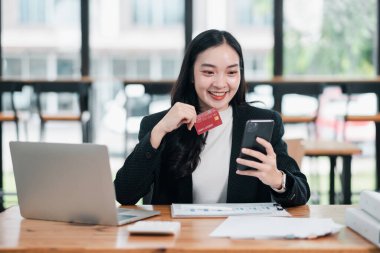 Parlak, modern ofis ortamında online alışveriş için akıllı telefon ve kredi kartı kullanan gülümseyen bir kadın..