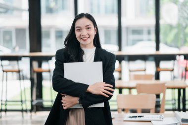 Genç profesyonel bir kadın, elinde dizüstü bilgisayarla, arka planda büyük pencereleri ve doğal ışığı olan modern bir ofiste güvenle duruyor..