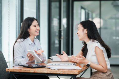 Modern ofis ortamında iki kadın, masadaki dijital cihazlar ve belgelerle çalışmayı tartışıyorlar..