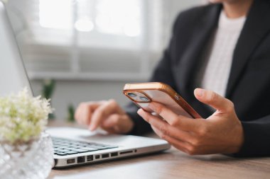 Modern ofis ortamında akıllı telefonu kontrol ederken dizüstü bilgisayarda çalışan bir profesyonel..