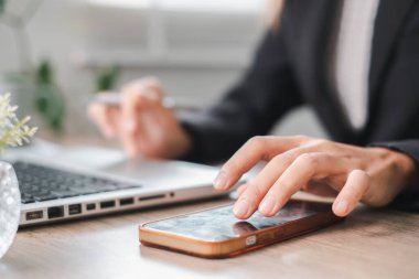 Modern çalışma ve iletişimi vurgulamak için bir masada akıllı telefon ve dizüstü bilgisayar kullanan bir profesyonelin yakın çekimi.