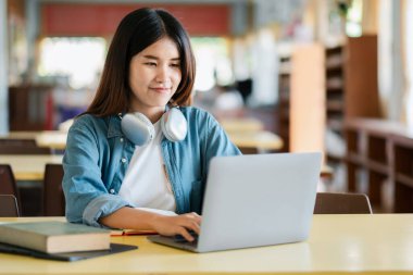 Kütüphanede kulaklıklı bir dizüstü bilgisayar kullanan genç bir kadın işe ya da çalışmaya odaklanmış, etrafı kitaplarla çevrili..