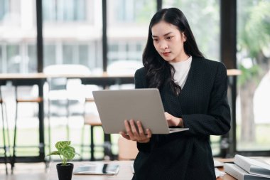 Profesyonel bir kadın, geniş pencereli ve doğal ışığı olan parlak, modern bir ofis ortamında dizüstü bilgisayar kullanıyor..