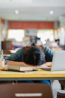 Yorgun bir öğrenci, akademik stres ve yorgunluğu betimleyen dizüstü bilgisayar ve kitaplarla çevrili bir masanın üzerinde dinleniyor..