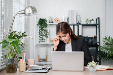 Modern bir ev ofisteki bir iş kadını, çevresi bitki ve ofis malzemeleriyle çevrili olarak dizüstü bilgisayarı üzerinde çalışırken stresli görünür..